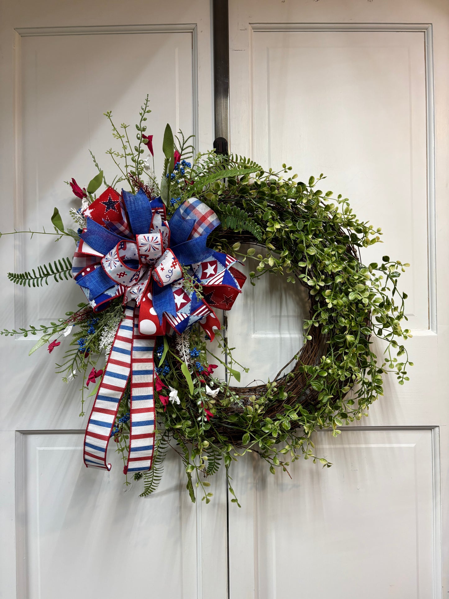 Patriotic greenery wreath featuring red, white, and blue decorations on a grapevine base, hanging on a door.
