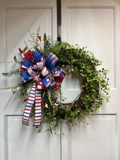 Patriotic greenery wreath featuring red, white, and blue decorations on a grapevine base, hanging on a door.