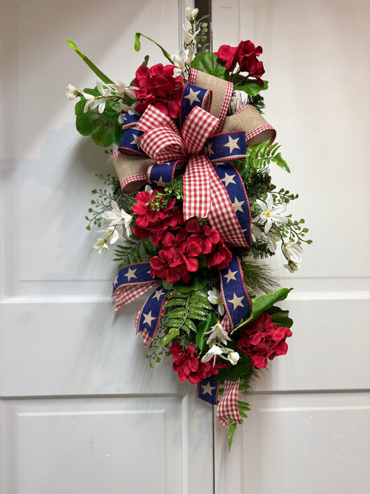 Patriotic geranium swag featuring red flowers, blue star ribbon, and white accents, hanging on a white door.