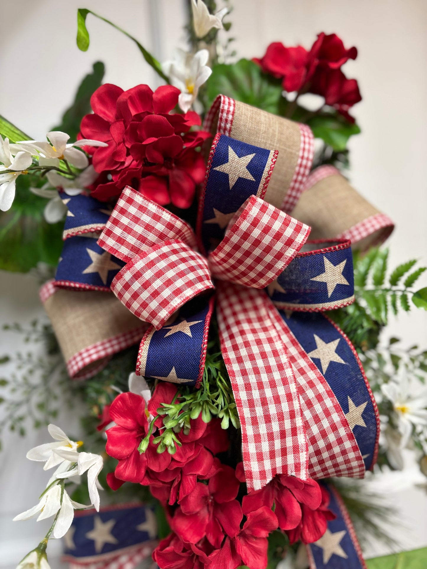 Patriotic geranium swag featuring red flowers, blue star-patterned ribbon, and a checkered bow on a teardrop design.